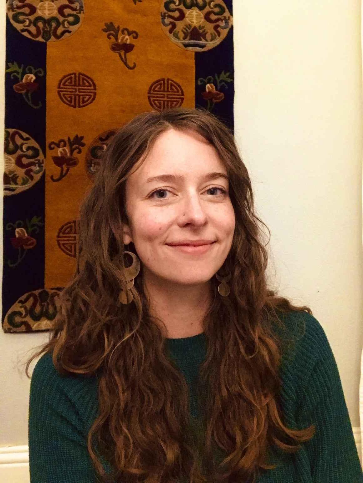 Kate Falkenhart, ACLU of Colorado operations manager, smiling, in front of light-colored wall with a brown and black tapestry hanging