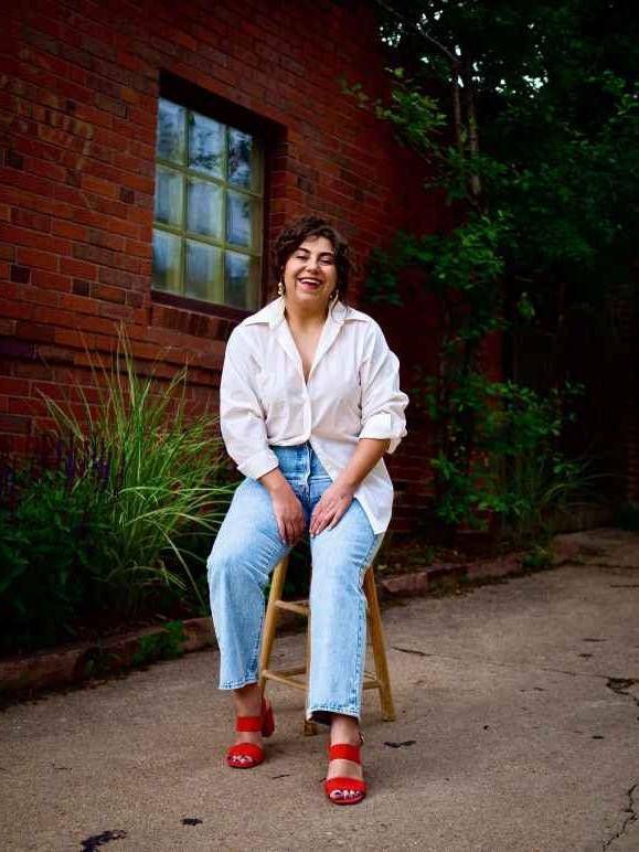 Marie Medina, smiling, outdoors, sitting on a stool in front of red brick building